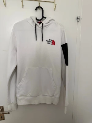 Vit hoodie från The North Face - Vit hoodie från The North Face med svart snörning i huvan och logotyp broderad på bröstet i svart och rött. Klassisk känguruficka framtill och ribbade muddar. Materialet är mjukt och bekvämt, perfekt för chill dagar.