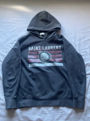 Mörkgrå Saint Laurent hoodie - Mörkgrå hoodie från Saint Laurent med stor huva och magficka. Tryck på bröstet med text och emblem i vitt och rött. Klassisk passform och ribbade muddar. Perfekt för dig som gillar streetstyle och vill ha något snyggt och bekvämt.