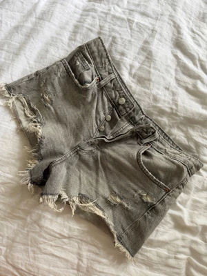 Grå slitna jeansshorts från Zara - Säljer ett par grå jeansshorts från Zara med slitna detaljer och fransiga kanter. Shortsen har hög midja, knappgylf och klassiska fem fickor. Perfekta för sommaren och ger en cool, avslappnad vibe.