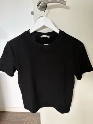 Svart t-shirt från Zara - En stilren svart t-shirt från Zara med rund halsringning och korta ärmar. T-shirten har en ribbad kant vid halsen och är tillverkad i ett mjukt material som känns skönt mot huden. Perfekt basplagg för alla tillfällen.