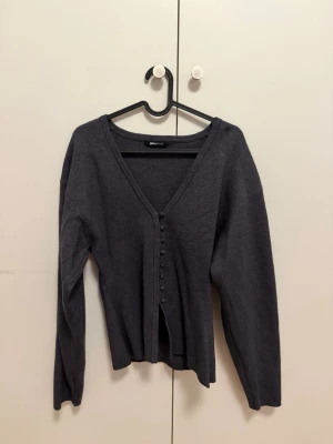 Mörkgrå kofta från Gina Tricot - Mörkgrå kofta från Gina Tricot med v-ringning och knappar framtill. Långärmad och i mjukt, tunt stickat material som ger en clean och enkel look. Perfekt att slänga över en topp eller klänning för extra lager.