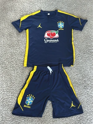 Brasilien fotbollsträningsset blå/gul - Snyggt fotbollsträningsset från Brasilien i marinblått med gula detaljer. Setet består av en t-shirt och shorts i lätt och ventilerande material. Både tröjan och shortsen har Brasil-logga och Jordan-logga, samt Guaraná Antarctica-tryck på bröstet. Perfekt för träning eller match!
