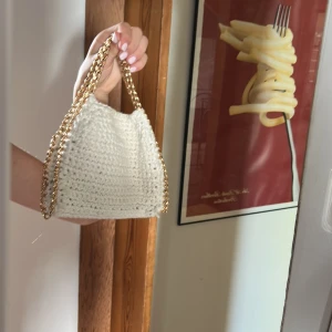 Vit stickad handväska med guldkedja - Superfin handväska i virkad stil, helt vit med chunky guldfärgad kedja som axelrem. Väskan har magnetlås och en mjuk, fyrkantig form. Perfekt för dig som vill ha en unik accessoar med trendig känsla. Liknar en stella Mc! Falabella storlek på pricken.