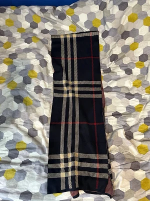 Mörkblå halsduk med rutigt mönster  - Snygg mörkblå halsduk med klassiskt rutigt mönster i beige, vitt och rött. Halsduken har en rektangulär form och är perfekt för att ge din outfit en stilren touch. Materialet känns mjukt och lent, troligen i ull eller akryl.