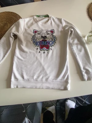 Vit sweatshirt med tiger från Kenzo - Säljer en vit sweatshirt från Kenzo med det ikoniska tigerhuvudet broderat i flera färger på bröstet och texten 'KENZO PARIS' under. Tröjan har rund halsringning, långa ärmar och är gjord i mjukt bomullsmaterial. Perfekt statement-plagg för dig som gillar streetwear.