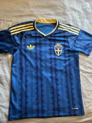 Blå Sverige fotbollströja  - Säljer en blå fotbollströja från Adidas med Sveriges landslagsemblem på bröstet och gula detaljer. Tröjan har klassiska Adidas-ränder på axlarna, gul krage och ett mönster i olika blå nyanser. Materialet är lätt och andas, perfekt för träning eller match.