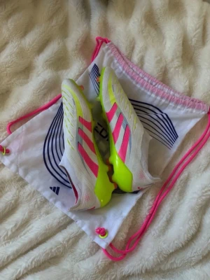 Adidas vita och neonrosa fotbollsskor - Säljer ett par snygga Adidas fotbollsskor i vitt med neonrosa och gula detaljer. Skorna har klassiska tre ränder på sidan, låg profil och greppvänlig sula för spel på gräs. Materialet är syntetiskt och designen är modern och sportig. Perfekta för dig som vill sticka ut på planen.