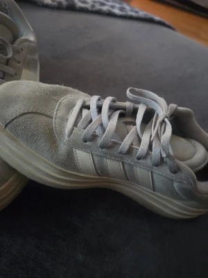 Gråa Adidas sneakers i mocka - Snygga grå sneakers från Adidas med ovandel i mocka och klassiska tre ränder på sidan. Skorna har rund tå, platt sula och matchande grå skosnören. Perfekta för en stilren och avslappnad look. Lite sliten men annars det bra ut.