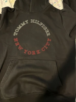 Svart hoodie Tommy Hilfiger NYC - Svart hoodie från Tommy Hilfiger med stor magficka och huva. På ryggen står det 'TOMMY HILFIGER' i vitt och 'NEW YORK CITY' i rött, båda i en cirkelformad design. Tillverkad i mjukt bomullsmaterial, perfekt för en avslappnad streetwear-look. ENDAST UPPHÄMTNING I STOCKHOLM 