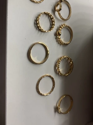 Set med åtta guldfärgade ringar - Säljer ett set med åtta tunna ringar i guldfärgad metall. Ringarna har olika mönster och texturer, vissa med räfflade kanter och små detaljer. Perfekta att mixa och matcha för en trendig look. Inget märke synligt.
