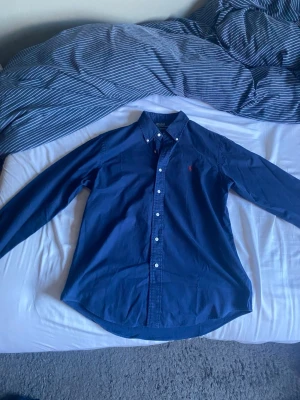 Marinblå skjorta från Polo Ralph Lauren - Säljer en klassisk marinblå skjorta från Polo Ralph Lauren med knappar hela vägen och den ikoniska röda loggan broderad på bröstet. Skjortan har lång ärm och button-down krage, perfekt till både jeans och chinos.