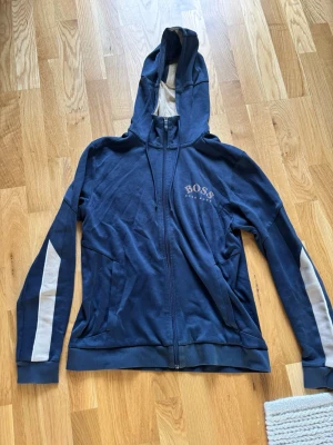 Blå hoodie från BOSS med dragkedja - Snygg blå hoodie från BOSS med logga på bröstet. Den har en liten slitning på höger armbåge ish, men är annars fläckfri. Storlek S