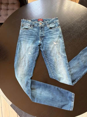 Jack&Jones jeans  - Säljer ett par blå jeans från jack&jones med slitningar och distressed detaljer. Jeansen har klassisk femficksmodell, knappgylf och smal passform. Perfekta för en avslappnad och trendig look.