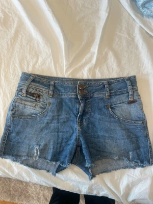 Blå jeansshorts med rå kant - Säljer ett par blå jeansshorts från Culture med råa kanter och slitningar framtill. Shortsen har klassisk femficksdesign, bälteshällor och dubbla knappar i midjan. Bakfickorna har snygga broderade detaljer i orange. Perfekta för sommaren!