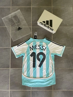Argentina Messi 19 fotbollströja Adidas - Säljer en klassisk Argentina fotbollströja från Adidas med Messi och nummer 19 på ryggen. Tröjan har ljusblå och vita vertikala ränder, svarta detaljer på axlarna och officiellt landslagsmärke på bröstet. Perfekt för fotbollsfans och samlare.