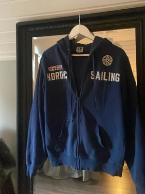 Blå hoodie från EP Collection - Mörkblå hoodie med dragkedja från EP Collection. Tryck med texten 'NORDIC SAILING' och flaggor på bröstet samt en kompassros. Utrustad med huva och fickor framtill. Storlek S, använd men i bra skick. 