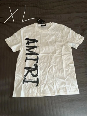 Vit AMIRI t-shirt med tryck - Säljer en vit t-shirt från AMIRI i storlek XL. T-shirten har ett stort svart vertikalt AMIRI-tryck på framsidan och är tillverkad i mjuk bomull. Klassisk passform med korta ärmar och rund hals. Perfekt för dig som gillar streetwear vibes.
