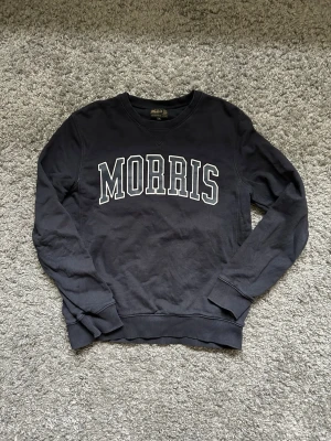 Mörkblå sweatshirt från Morris - Klassisk mörkblå sweatshirt från Morris med vit text på bröstet. Tröjan har rund halsringning, ribbade muddar vid ärmslut och nederkant samt en avslappnad passform. Perfekt för en casual och sportig stil.