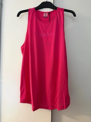 Rosa träningstopp från Craft - Snyggt rosa träningslinne från Craft med rund halsringning och diskret logga framtill. Tillverkat i ett lätt och ventilerande funktionsmaterial som passar perfekt för gymmet eller löprundan.