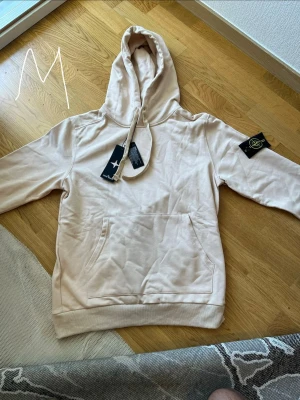 Beige hoodie från Stone Island - Snygg beige hoodie från Stone Island med klassisk patch på vänster ärm och stor känguruficka framtill. Hoodien har dragsko i huvan och ribbade muddar vid ärmslut och nederkant. Tillverkad i mjuk bomull för skön känsla.