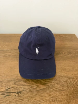 Mörkblå keps från Polo Ralph Lauren - Snygg mörkblå keps från Polo Ralph Lauren med klassisk vit broderad logga framtill och 'POLO' broderat bak. Justerbart spänne i metall baktill för perfekt passform. Tillverkad i bomullstyg som andas.