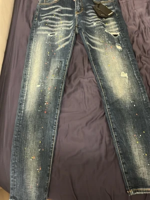 Mörkblå jeans med färgstänk dsq2 - Säljer ett par mörkblå jeans med slitningar och färgstänk i olika färger för en cool och unik look. Jeansen har raka ben, klassiska fem fickor och kontrastsömmar. Perfekta för dig som vill sticka ut med din stil.