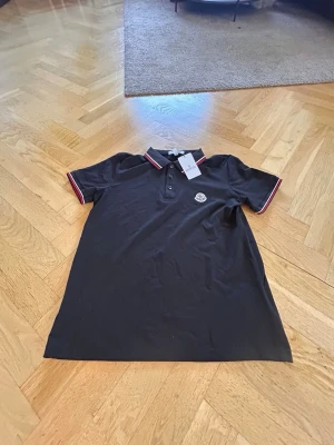 Moncler Piké  - Klassisk svart pikétröja från Moncler med korta ärmar och rödvita detaljer på krage och ärmslut. Tröjan har knappslå framtill och Moncler-logga på bröstet. Tillverkad i mjuk bomull för en bekväm känsla.