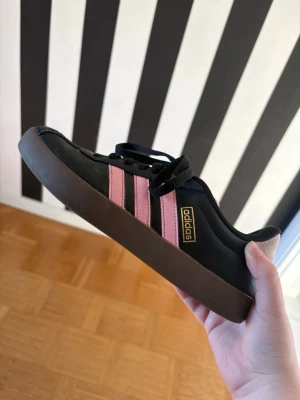 Svarta Adidas sneakers med rosa ränder - Snygga svarta Adidas sneakers med klassiska rosa ränder på sidorna och brun sula. Skorna har lågt skaft, svart snörning och är tillverkade i slitstarkt material. Perfekta för dig som vill ha en stilren och sportig look.