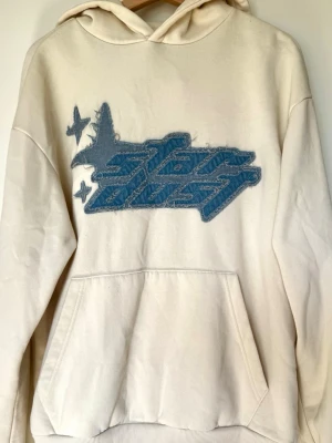 Beige hoodie med blått Star Dust-jeanstryck - Beige hoodie med stor blå jeanstryck framtill där det står ”Star Dust.” Hoodien har en avslappnad passform och kommer från det polska märket House som fokuserar på streetwear. Kvaliteten är bra och har bara fåtal tecken på användning som väldigt små märken på tyget. Storleken är en S i herr och är oversized. Jag tycker hoodien ger lite 2000’s Y2K känsla. 