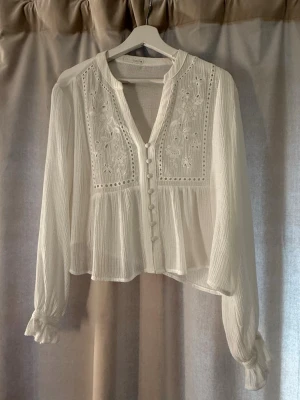 Vit blus med broderi och knappar - Romantisk vit blus med långa ärmar, broderade blommor på bröstet och klädda knappar framtill. Tillverkad i ett tunt, lätt och något transparent tyg med struktur. Blusen har volangdetaljer vid ärmslut och en avslappnad passform🤍🤍🤍