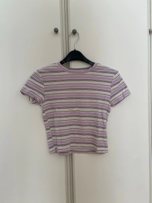 Ljuslila randig t-shirt - Säljer en t-shirt i ljuslila och vitt med tunna svarta ränder. T-shirten har korta ärmar, rund halsringning och är ribbad i materialet. Det är barnstorleken 146/152 cm.