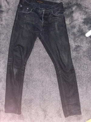 Svarta nudie jeans - Säljer ett par svarta nudie jeans med snygga detaljer och fadeing. Passformen är frealess freddie och är i storlek W30 L34. Perfekta för en trendig och avslappnad stil. 