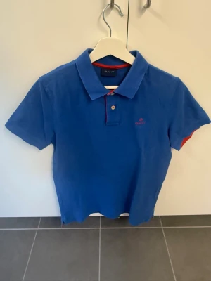 Blå pikétröja från Gant - Snygg blå pikétröja från Gant med klassisk krage och korta ärmar. Tröjan har röda detaljer vid knäppningen och Gant-logga broderad på bröstet. Perfekt till jeans eller shorts för en avslappnad stil.