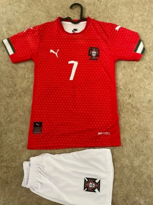 Portugal Ronaldo matchställ Puma - Säljer ett rött Portugal matchställ med Ronaldo 7-tryck. Tröjan har vita detaljer, landslagets emblem och Puma-logga. Korta ärmar med gröna och vita ränder vid ärmslut. Tillhörande vita shorts med emblem. Materialet är lätt och andas, perfekt för fotboll.