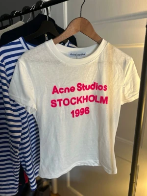 Vit Acne Studios t-shirt med tryck - Vit t-shirt från Acne Studios med rosa texttryck 'Acne Studios STOCKHOLM 1996' på bröstet. Klassisk rund halsringning och korta ärmar. Perfekt för en trendig och avslappnad stil.