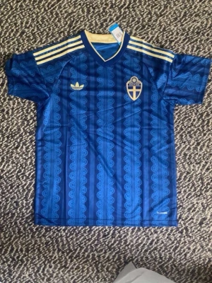Blå Sverige fotbollströja Adidas - Säljer en blå fotbollströja från Adidas med Sveriges landslagsemblem på bröstet. Tröjan har guldfärgade ränder på axlarna, mönstrad design och klassisk Adidas-logga. Perfekt för dig som vill stötta Sverige med stil.