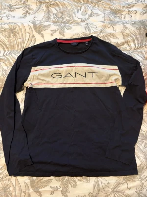Mörkblå långärmad topp från GANT - Snygg mörkblå långärmad topp från GANT med beige panel och röda detaljer över bröstet. Stor GANT-logga framtill. Tillverkad i mjuk bomull och har en klassisk passform. Perfekt för dig som gillar stilrena och bekväma plagg.