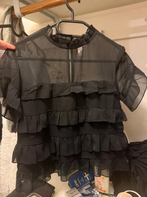Svart volangblus med transparent tyg - Säljer en svart blus med flera lager volanger och transparent tyg. Blusen har korta ärmar och hög hals, vilket ger en trendig och luftig look. Perfekt för dig som gillar statement-plagg med mycket detaljer.