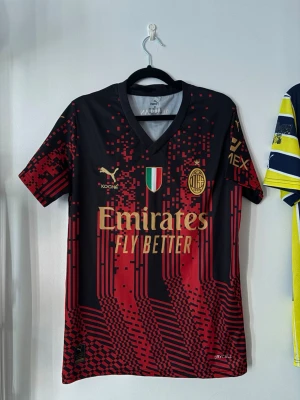 AC Milan matchtröja Puma svart/röd - Snygg AC Milan matchtröja från Puma i svart och rött med digitalt pixelmönster. Tröjan har guldfärgade loggor, V-ringning och korta ärmar. Materialet är funktionspolyester (Drycell) som andas. Perfekt för fotbollsträning eller att bära till vardags.