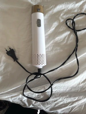 Multistyler för hår med flera munstycken - Säljer en elektrisk multistyler för hår i vitt och guld med flera utbytbara munstycken, inklusive rundborste, locktillbehör och fönmunstycke. Perfekt för att skapa olika frisyrer hemma. Drivs med sladd och har enkel knappkontroll.
