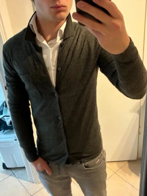 Mörkgrå cardigan med krage från WE - Stilren mörkgrå cardigan från WE med klassisk krage och knappstängning framtill. Tröjan har en bröstficka och är tillverkad i mjukt, tunt material som passar perfekt för lager på lager. Långärmad och enkel att matcha till olika outfits.