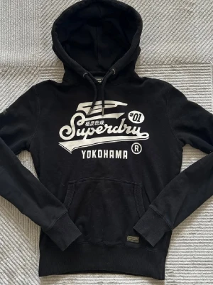 Svart Superdry hoodie med tryck - Svart hoodie från Superdry med stort vitt tryck på bröstet där det står 'Superdry Yokohama'. Klassisk känguruficka framtill och justerbar huva med dragsko. Perfekt för en avslappnad streetstyle-look.