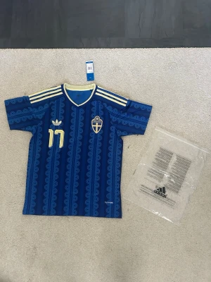 Blå Sverige fotbollströja Adidas - Säljer en blå Sverige fotbollströja från Adidas med gula detaljer, nummer 17 och namnet Gyökeres på ryggen. Tröjan har ett mönstrat tryck, korta ärmar och klassiska Adidas-ränder på axlarna. Perfekt för dig som vill stötta landslaget med stil.