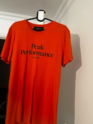 Orange Peak Performance t-shirt - Snygg orange t-shirt från Peak Performance med svart tryck framtill. Klassisk rund halsringning och korta ärmar. Perfekt för en avslappnad och sportig stil.