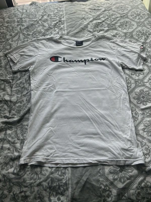 Vit Champion t-shirt med logga - Säljer en klassisk vit t-shirt från Champion i storlek XL. T-shirten har korta ärmar och den ikoniska Champion-loggan tryckt över bröstet. Tillverkad i mjuk bomull och har en avslappnad passform. Perfekt till alla tillfällen där du vill ha en clean och sportig look.