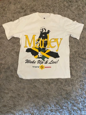 Vit Marley t-shirt  - Säljer en vit t-shirt från Pull&Bear med stort Marley-tryck framtill och texten 'Wake Up & Live!'. T-shirten har klassisk passform, rund hals och är kortärmad. Materialet är mjuk bomull och trycket har inslag av gult, grönt och svart. Storlek S men mer som än M