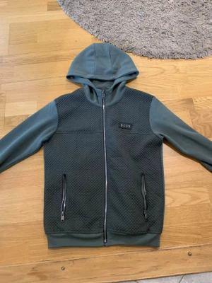 Grön hoodie-cardigan från BOSS - Stilren grön hoodie-cardigan från BOSS med dragkedja framtill och två fickor med dragkedja. Framsidan har ett quiltat mönster och ärmarna samt huvan är i mjukt tyg. BOSS-logga på bröstet och detaljer i metall. Perfekt för en avslappnad men snygg look.