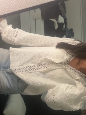 Vit blus med spetsdetaljer - Superfin vit blus med långa ärmar och romantiska spetsdetaljer längs ärmslut och halsringning. Blusen har en lätt rynkad struktur och knytning framtill. Perfekt att styla med jeans eller kjol! 