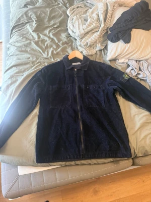 Mörkblå overshirt från Stone Island - Säljer en mörkblå overshirt från Stone Island i manchestertyg. Jackan har två bröstfickor, dragkedja framtill och klassisk krage. Ikonisk logotyp på vänster ärm. Perfekt för lager-på-lager och streetwear-stil.
