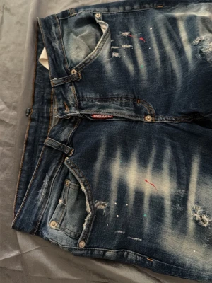 Dsquared2 slitna blå jeans - Säljer ett par blå jeans från Dsquared2 med slitningar, blekta partier och färgstänk för en riktigt edgy look. Klassisk femficksmodell med knappgylf och detaljerade sömmar. Perfekta för dig som gillar streetstyle och vill sticka ut.